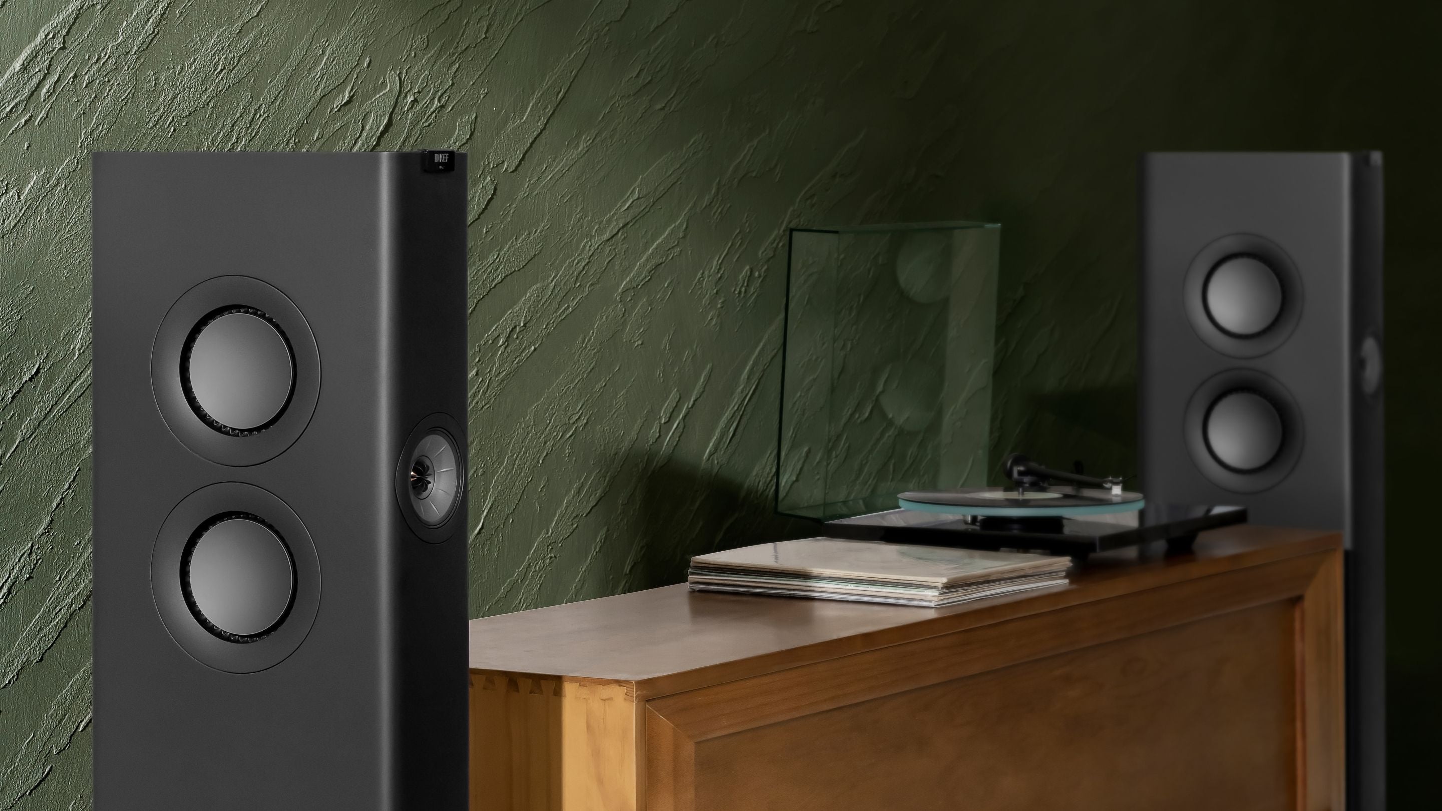 Pair of LS60 Wireless black speakers next to a wooden surface with a green textured wall in the background