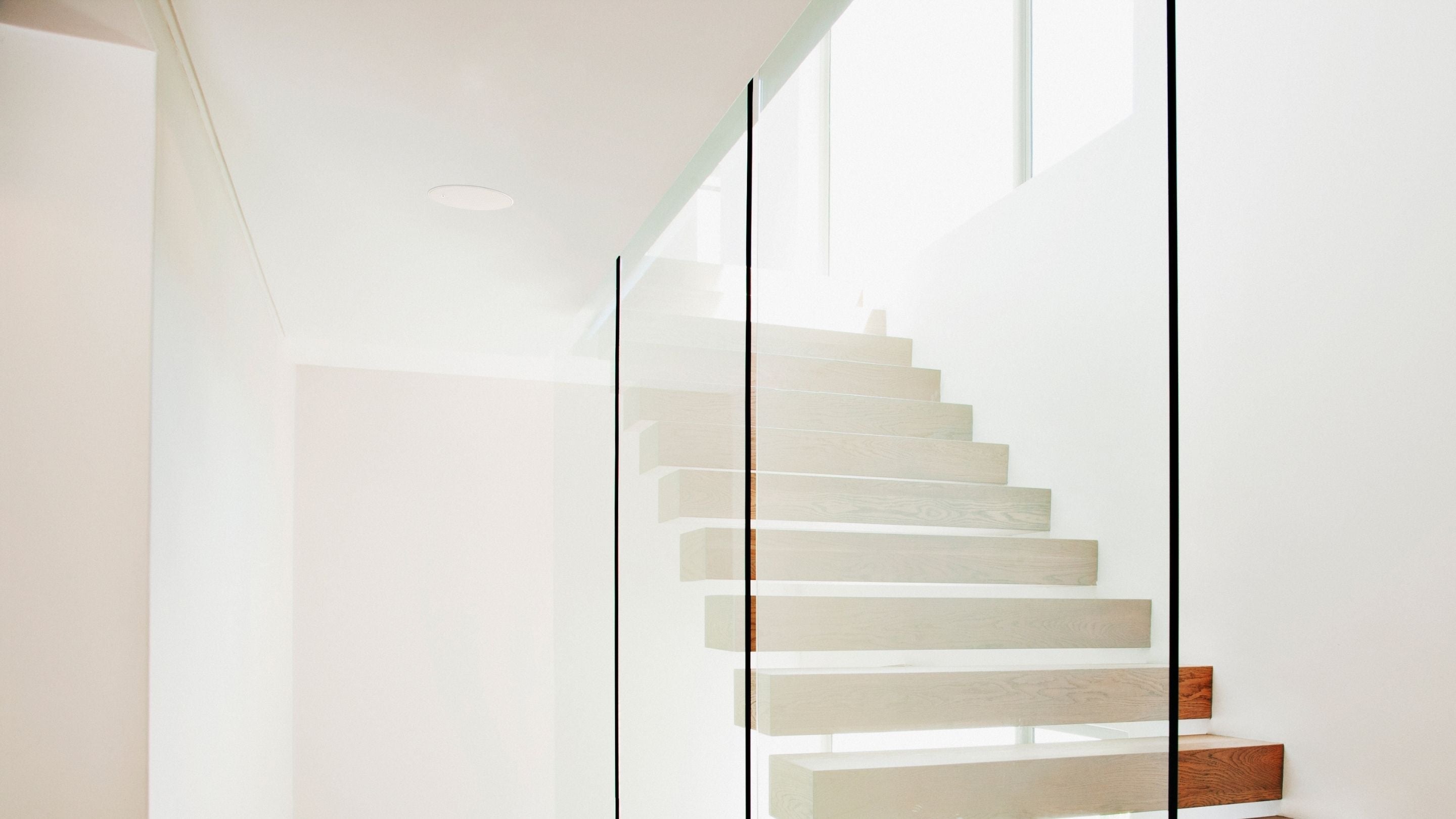 KEF architectural speakers displayed alongside a sleek glass stairway with wooden steps in a minimalist white room.
